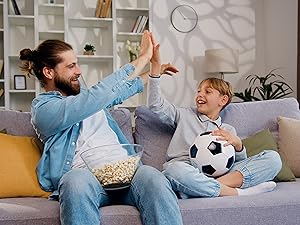 Man and boy on couch, celebrating while watching sports. Man holds popcorn bowl, boy holds soccer ball. Living room setting with bookshelves.