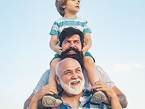 Three generations of males outdoors against a blue sky. A young boy sits on the shoulders of a bearded man, who is on the shoulders of an older man with a white beard.