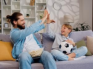 Man and boy on couch, celebrating while watching sports. Man holds popcorn bowl, boy holds soccer ball. Living room setting with bookshelves.