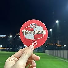 Hand holding a round, red paper fan with 'Thierry Henry' printed on it. Nighttime stadium setting visible in the background.