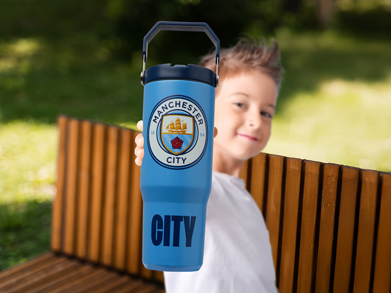 Text visible: 'CITY' and Manchester City Football Club logo. Blue sports water bottle with club crest against outdoor park bench background.