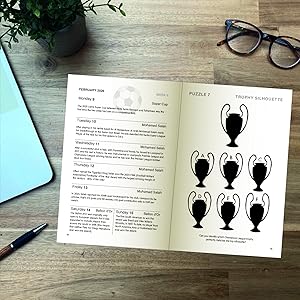 Open notebook showing text page and illustration page with six trophy designs. Eyeglasses and blue device visible on wooden desk surface.
