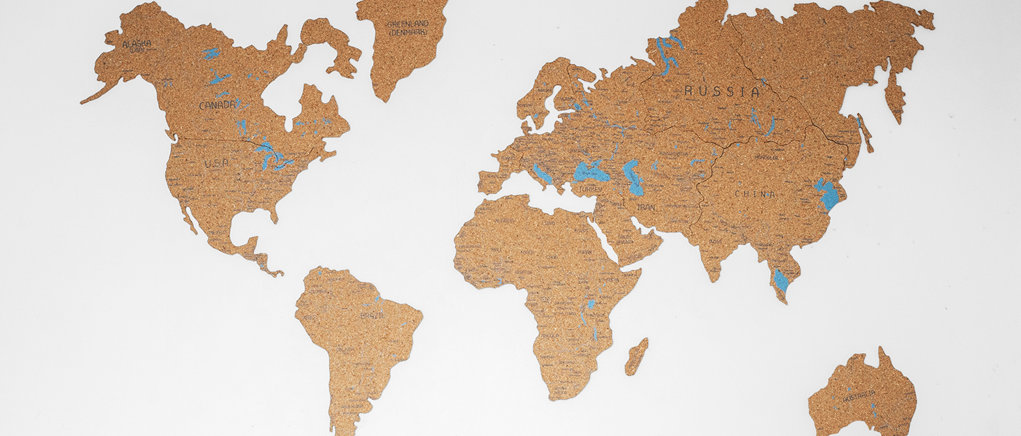 Cork board world map with continents outlined in brown cork material. Small blue pins mark various locations across different continents.